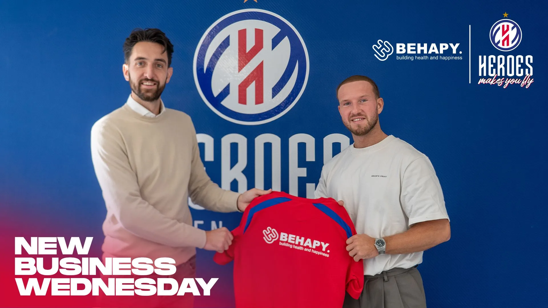 Twee mannen houden samen het rode Heroes inloopshirt omhoog met het BEHAPY-logo op de achterkant, voor de clubbanner van Heroes Den Bosch.